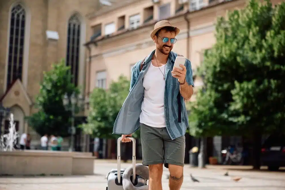 A male tourist pulling his luggage behind him and looking at his phone.